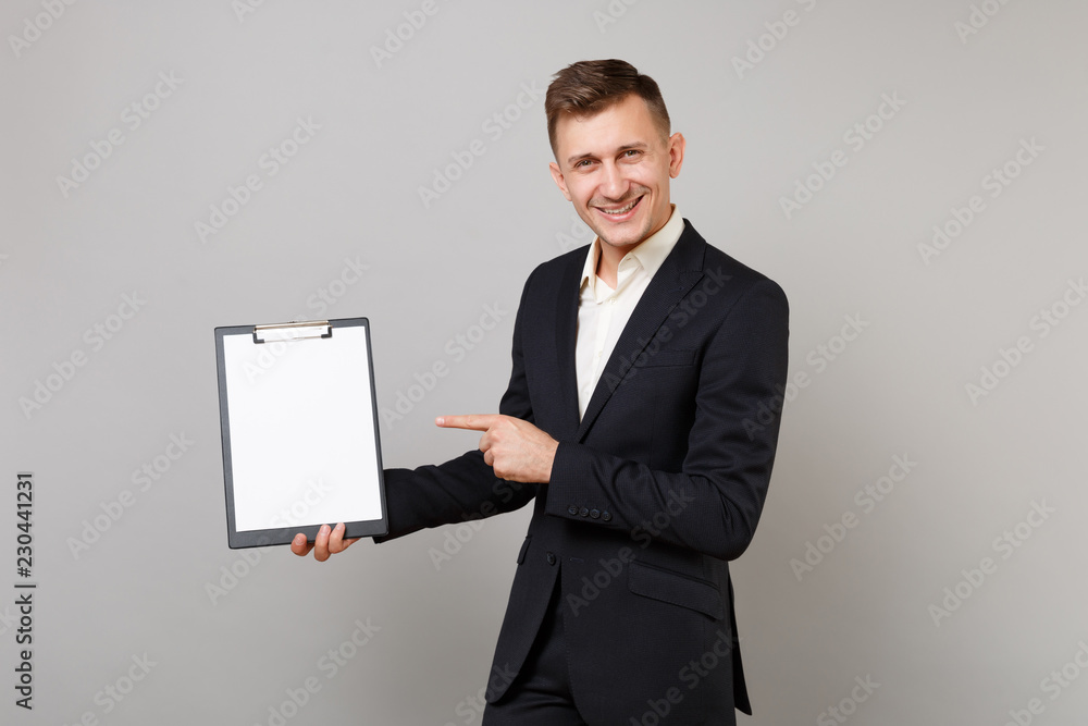 Smiling young business man in suit pointing index finger on clipboard with blank empty sheet workspace isolated on grey wall background. Achievement career wealth business concept. Mock up copy space. Smiling young business man in suit pointing index finger on clipboard with blank empty sheet workspace isolated on grey wall background. Achievement career wealth business concept. Mock up copy space.