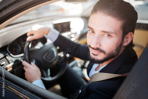 Satisfied and happy man sitting in car and posing. He looks at camera and smiles a bit. Bearded guy holds hands on steering wheel.