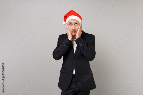 Tired upset young business man in Christmas hat looking up, put hands on cheeks isolated on grey background. Achievement career wealth business. Happy New Year 2019 celebration holiday party concept. © ViDi Studio