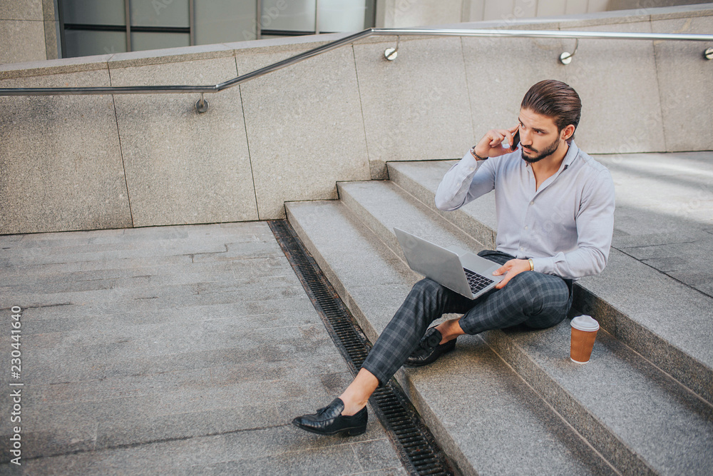 Picture of young businessman sits on steps and works. He talks on phone and looks at laptop's screen. Young man keeps his legs crossed.