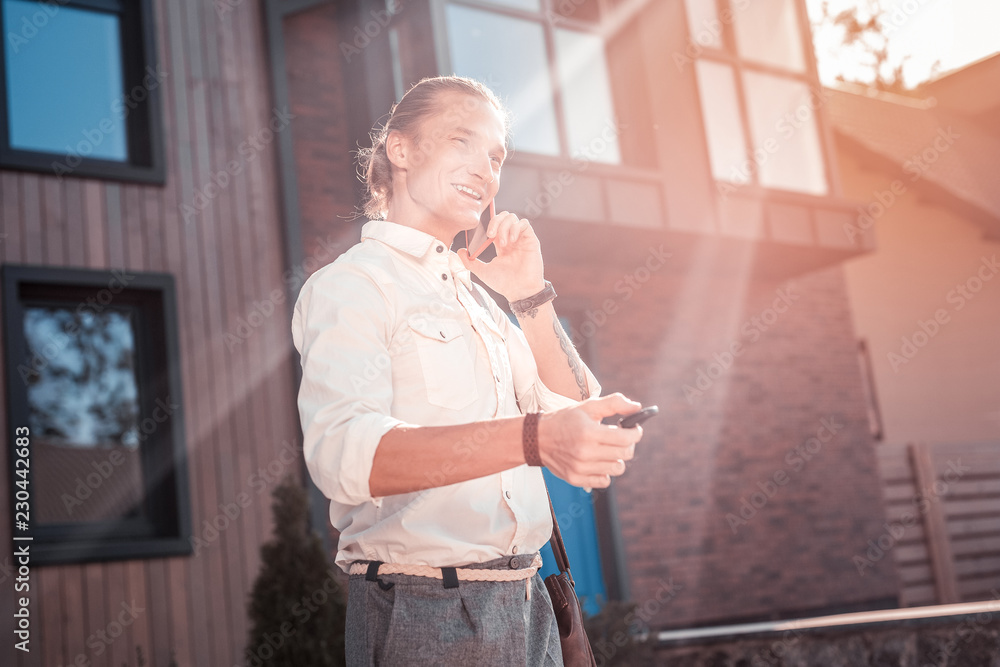 Fototapeta premium Cheerful man. Handsome businessman feeling cheerful while calling his wife on way to work and opening his car