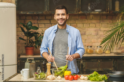 It's so delicious! Casual happy young man preparing salad at home in loft kitchen and smiling.