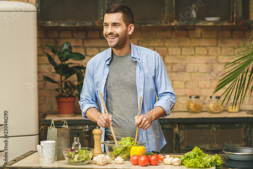 It's so delicious! Casual happy young man preparing salad at home in loft kitchen and smiling.