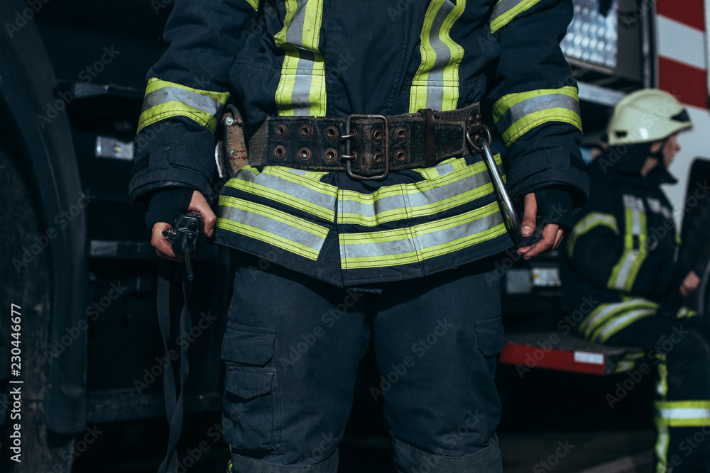 cropped shot of firefighter in fireproof uniform and colleague behind at fire station cropped shot of firefighter in fireproof uniform and colleague behind at fire station
