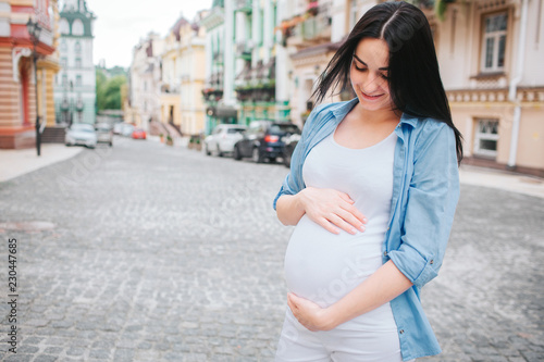 Portrait of a happy black hair and proud pregnant woman in a city in the background. Photo of female model touching her belly with hands.