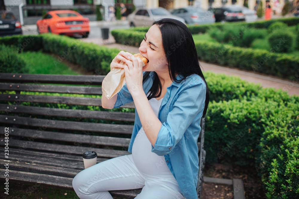 Fototapeta premium Portrait of a happy black hair and proud pregnant woman in a city in the background. Hungry expectant mother is listening to music in the park with an unborn child and eating a pie or sandwich. Hungry