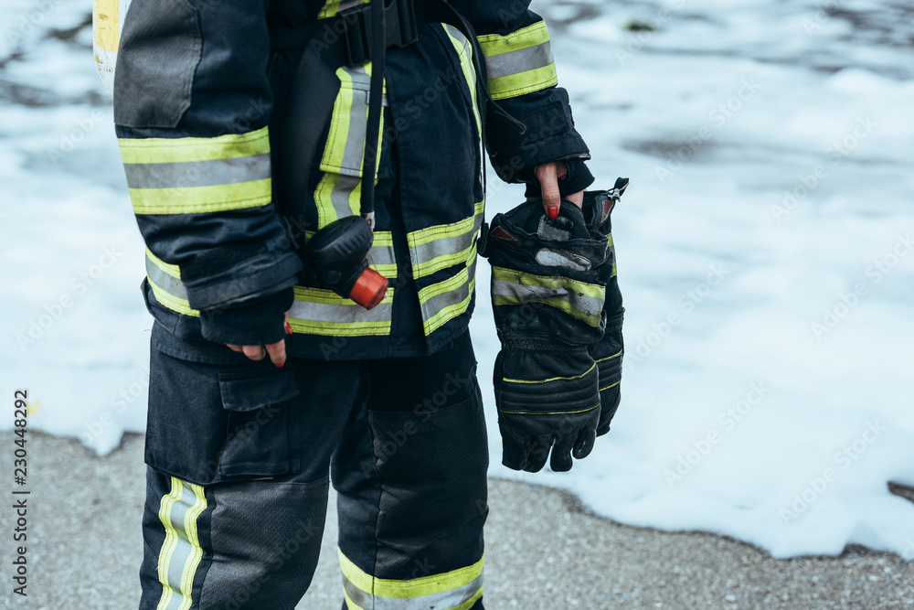 partial view of female firefighter in fireproof uniform with gloves in hand standing on steet with foam partial view of female firefighter in fireproof uniform with gloves in hand standing on steet with foam