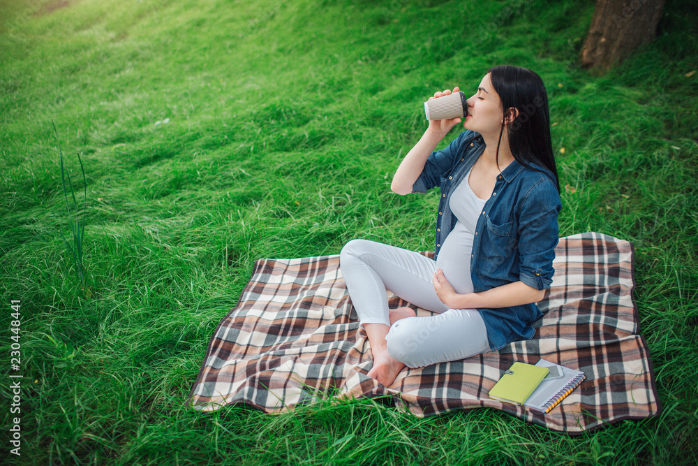 Portrait of a happy black hair and proud pregnant woman in a city in the park . Photo of female model touching her belly with hands. The female model is sitting on grass and drinking coffee or tea. Portrait of a happy black hair and proud pregnant woman in a city in the park . Photo of female model touching her belly with hands. The female model is sitting on grass and drinking coffee or tea.