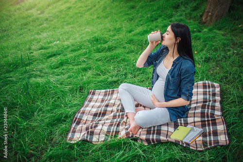 Portrait of a happy black hair and proud pregnant woman in a city in the park . Photo of female model touching her belly with hands. The female model is sitting on grass and drinking coffee or tea.