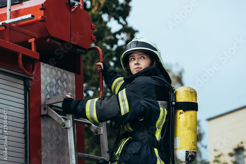 Fotografija female firefighter with fire extinguisher on back standing on fire truck on stre