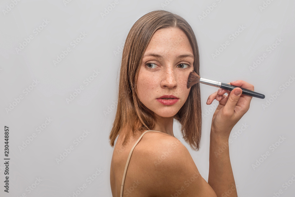 Beautiful and nice young woman stands and poses. She puts make up with brush on her chicks. She looks to left and turns behind. Isolated on grey background. Beautiful and nice young woman stands and poses. She puts make up with brush on her chicks. She looks to left and turns behind. Isolated on grey background.
