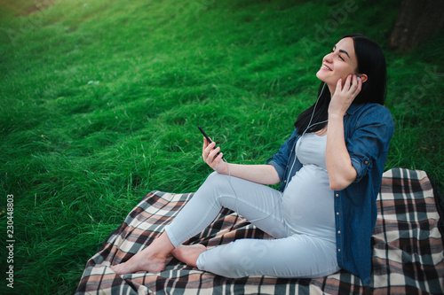 Portrait of a happy black hair and proud pregnant woman in a city in the background. She is sitting on a city bench. Expectant mother is listening to music in the park with an unborn child