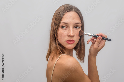 Beautiful and nice young woman stands and poses. She puts make up with brush on her chicks. She looks to left and turns behind. Isolated on grey background.