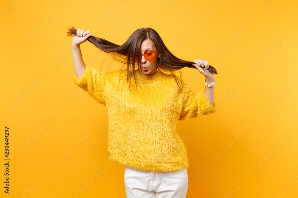 Pretty young woman in fur sweater, heart orange glasses fool around, holding her hair like ponytails isolated on bright yellow background. People sincere emotions, lifestyle concept. Advertising area.