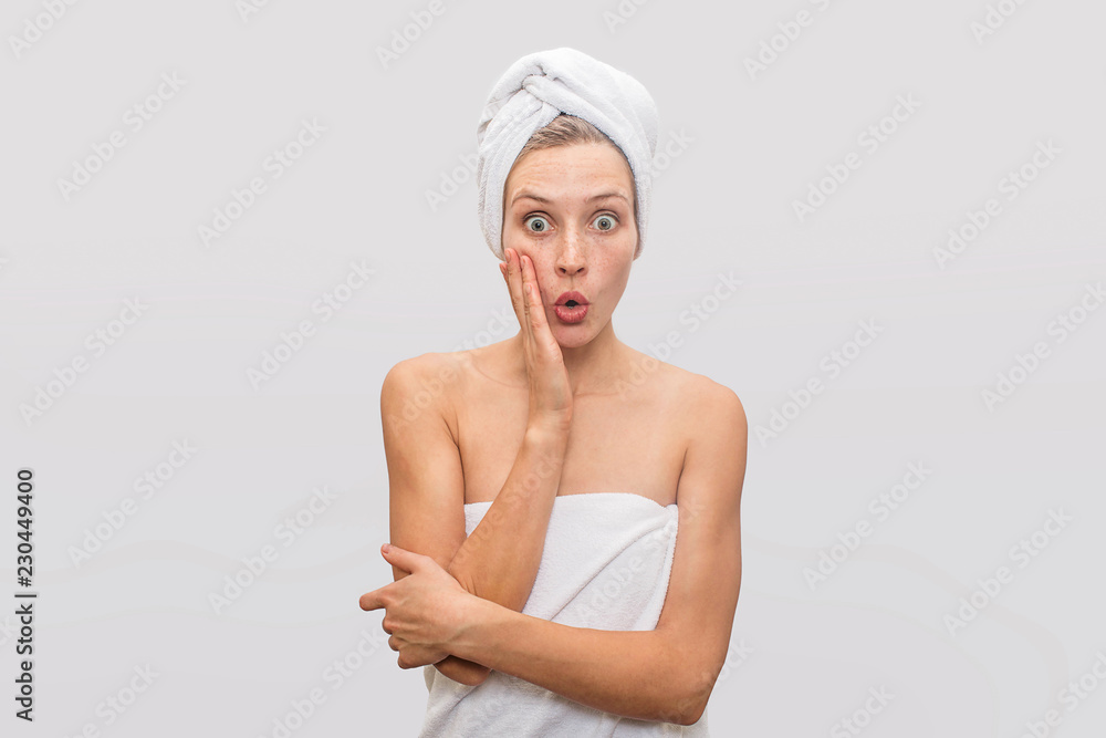 Beautiful and wondered young freckled woman stands and poses on camera. She is excited. Her body and hair are covered with white towels. Isolated on grey background.