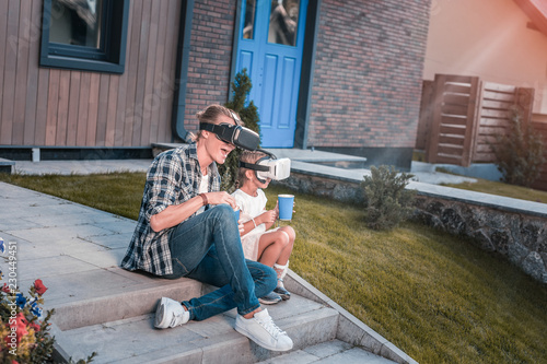 Eating popcorn. Modern father and daughter wearing virtual reality glasses and eating popcorn sitting on the porch