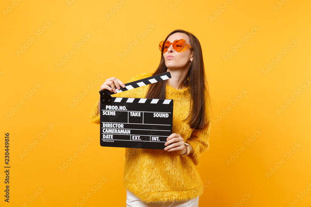 Beautiful young woman in orange heart eyeglasses looking aside and holding classic black film making clapperboard isolated on yellow background. People sincere emotions, lifestyle. Advertising area.