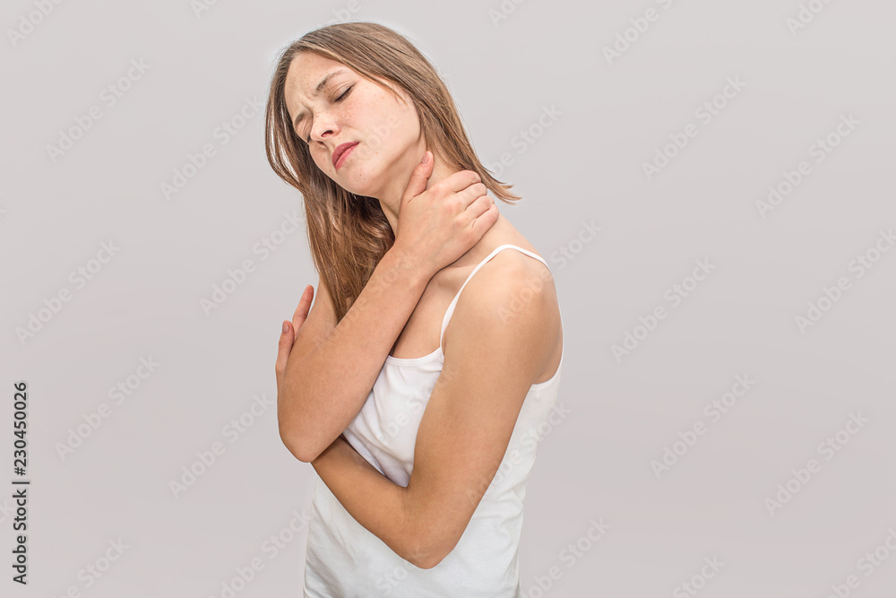 Naklejka premium Young woman feels pain in neck. She keeps eyes closed and hold hands together. Model wears white t-shirt. Isolated on grey background.