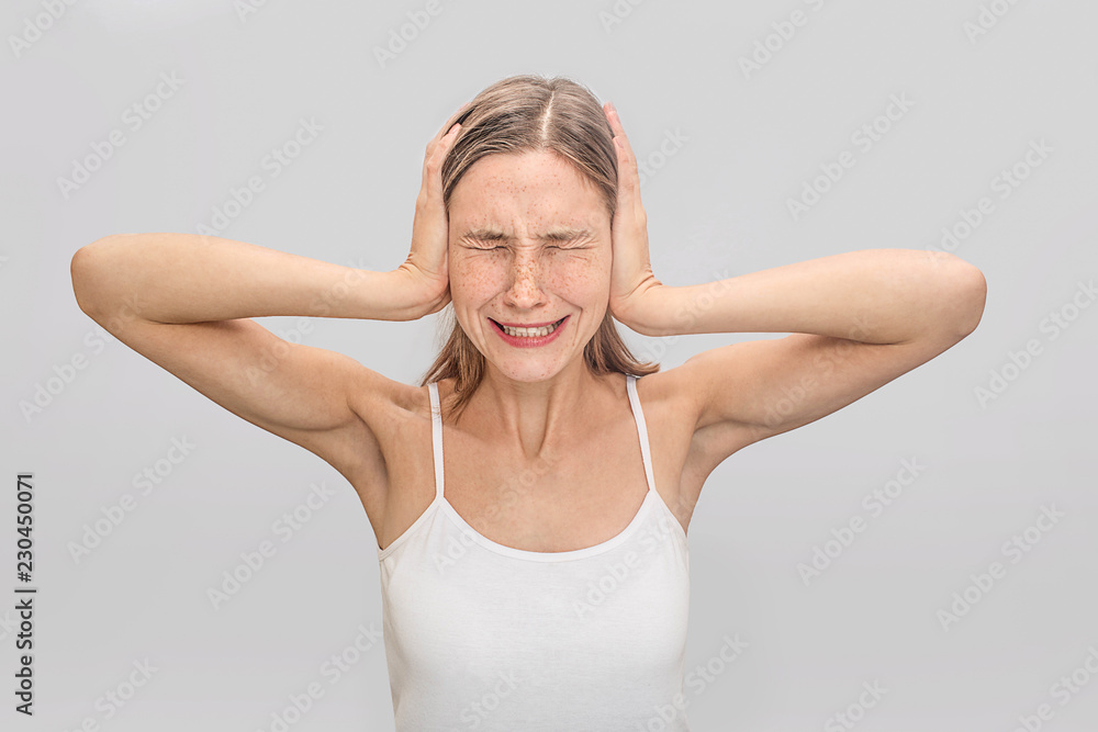 Young woman suffers from headache. She coveres ears with hands and keeps eyes closed. Young woman wears white t-shirt. Isolated on grey background. Young woman suffers from headache. She coveres ears with hands and keeps eyes closed. Young woman wears white t-shirt. Isolated on grey background.