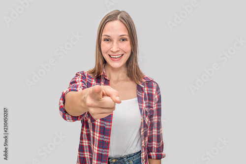 Nice and positive young woman stands and points on camera. She smiles. Model wears white t-shirt and cello shirt. Isolated on grey background.