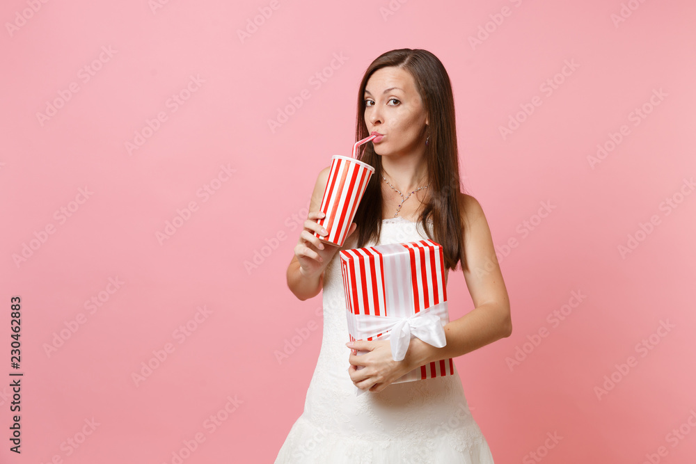 Portrait of indifferent bride woman in wedding dress hold red box with gift, present drinking cola or soda from plastic cup isolated on pink pastel background. Wedding celebration concept. Copy space. Portrait of indifferent bride woman in wedding dress hold red box with gift, present drinking cola or soda from plastic cup isolated on pink pastel background. Wedding celebration concept. Copy space.