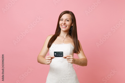 Portrait of smiling beautiful bride woman in white wedding dress holding credit card isolated on pink pastel background. Organization of wedding celebration concept. Copy space for advertisement.