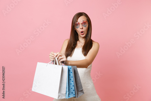 Shocked pretty bride woman in white wedding dress and heart glasses holding multi colored packages bags with purchases after shopping isolated on pink background. Organization of wedding. Copy space.