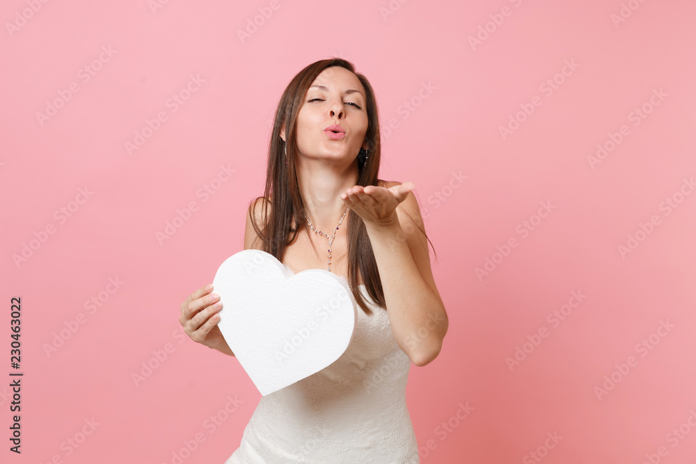 Tender bride woman with closed eyes in wedding dress blowing lips, send air kiss, holding white heart with copy space isolated on pastel pink background. Wedding celebration concept. Advertising area. Tender bride woman with closed eyes in wedding dress blowing lips, send air kiss, holding white heart with copy space isolated on pastel pink background. Wedding celebration concept. Advertising area.