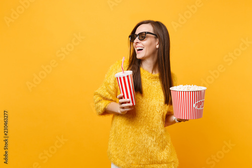 Joyful young woman in 3d imax glasses looking aside watching movie film hold bucket of popcorn, plastic cup of cola or soda isolated on yellow background. People sincere emotions in cinema, lifestyle.