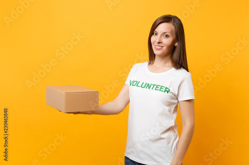 Portrait of smiling woman in white t-shirt with written inscription green title volunteer with blank cardboard box isolated on yellow background. Voluntary free assistance help, charity grace concept.