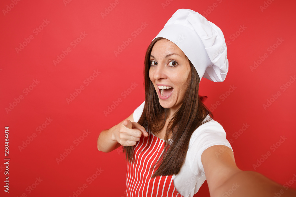 Housewife female chef cook or baker in striped apron, white t-shirt, toque chefs hat isolated on red wall background. Close up housekeeper woman doing taking selfie shot. Mock up copy space concept. Housewife female chef cook or baker in striped apron, white t-shirt, toque chefs hat isolated on red wall background. Close up housekeeper woman doing taking selfie shot. Mock up copy space concept.