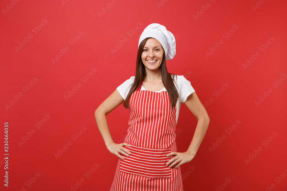 Housewife female chef cook or baker in striped apron, white t-shirt, toque chefs hat isolated on red wall background. Beautiful housekeeper woman standing with arms akimbo. Mock up copy space concept. Housewife female chef cook or baker in striped apron, white t-shirt, toque chefs hat isolated on red wall background. Beautiful housekeeper woman standing with arms akimbo. Mock up copy space concept.