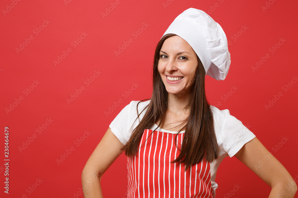 Housewife female chef cook or baker in red striped apron, white t-shirt, toque chefs hat isolated on red wall background. Close up portrait of housekeeper brunette woman. Mock up copy space concept. Housewife female chef cook or baker in red striped apron, white t-shirt, toque chefs hat isolated on red wall background. Close up portrait of housekeeper brunette woman. Mock up copy space concept.