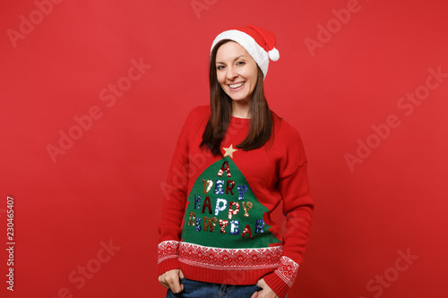 Smiling pretty young Santa girl in knitted sweater, Christmas hat holding hands in pockets isolated on bright red background. Happy New Year 2019 celebration holiday party concept. Mock up copy space. photo
