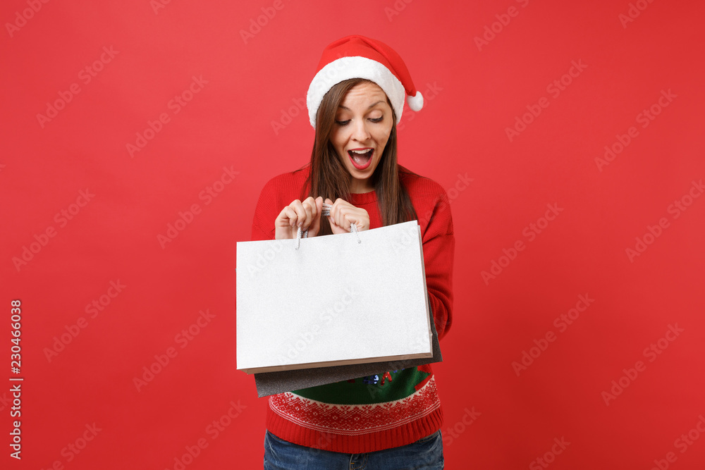 Surprised curious young Santa girl keeping mouth wide open, looking on packages bags with purchases after shopping isolated on red background. Happy New Year 2019 celebration holiday party concept. Surprised curious young Santa girl keeping mouth wide open, looking on packages bags with purchases after shopping isolated on red background. Happy New Year 2019 celebration holiday party concept.