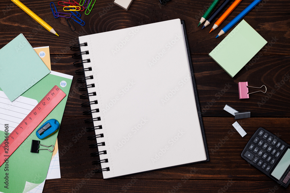 Empty spiral notebook with white pages and multicolored stationery on a brown wooden table, top view. Back to school concept. Empty spiral notebook with white pages and multicolored stationery on a brown wooden table, top view. Back to school concept.