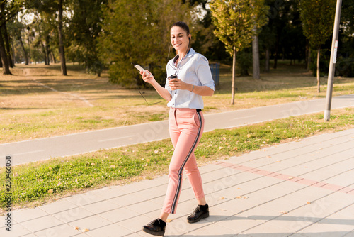 Happy young woman with coffee listening music on smartphone and walking on promenade © BGStock72