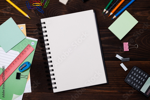 Empty spiral notebook with white pages and multicolored stationery on a brown wooden table, top view. Back to school concept.