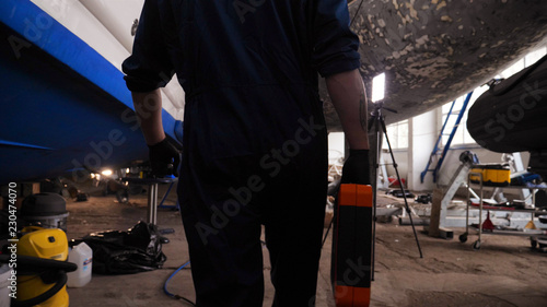 A young handsome professional engineer (mechanic) walks through the garage of the yachts with a tools in his hands. Concept of: Work start, Work class, Yacht club, Service, Garage, Tools, Work Clothes