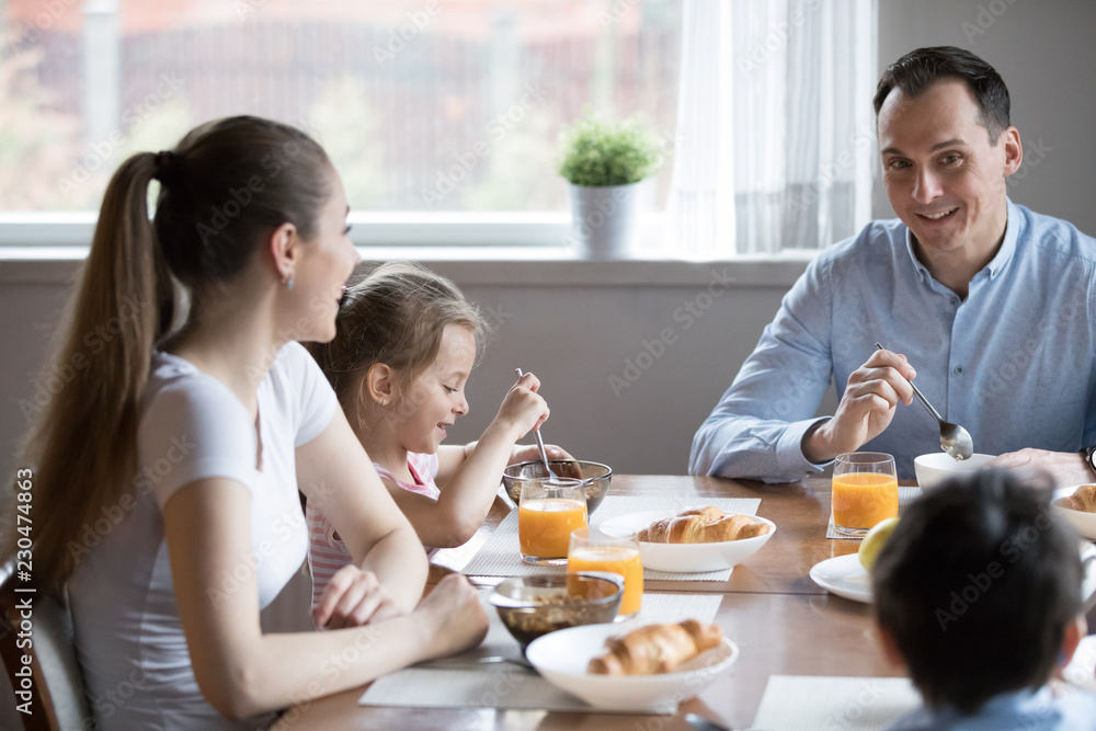 Attractive positive young family with little adorable preschool children sitting together in kitchen in the morning at home having breakfast eating healthy food feels good happy talking to each other.