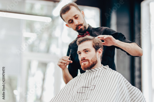 Stylish mustachioed barber dressed in a black shirt with a red bow tie scissors the hair of a young man in a barbershop