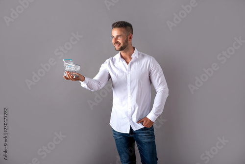 A young man in a white shirt holding a mini shopping cart and standing in front of a grey background in the studio.