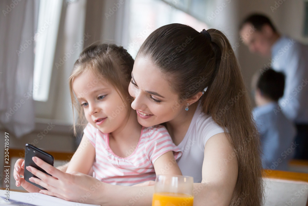 Woman have fun together with little adorable girl sitting indoors. Mother and daughter looking at mobile phone screen watching cartoons making video call or playing games father with son on background Woman have fun together with little adorable girl sitting indoors. Mother and daughter looking at mobile phone screen watching cartoons making video call or playing games father with son on background