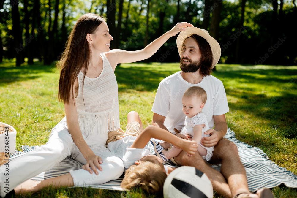 Warm sunny day in the park. Stylish young family dressed in white casual clothes is sitting on a blanket on the green grass.