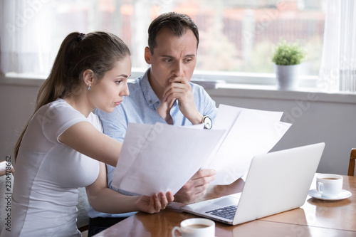 Stressed millennial married couple sitting at the desk at home use laptop checking documents unpaid bills, taxes, due debt, bank account balance. Bankruptcy lack of money financial problems concept