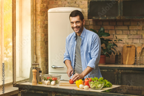 It's so delicious! Casual happy young man preparing salad at home in loft kitchen and smiling, using laptop.