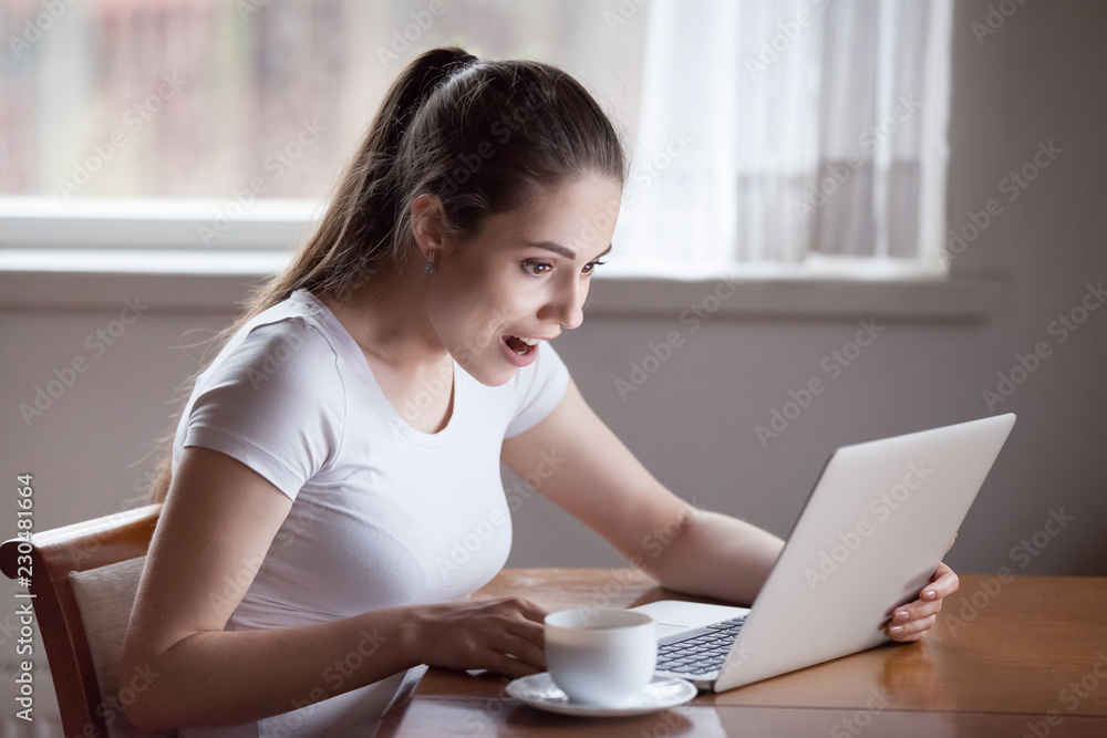 Young positive surprised girl sitting in kitchen at table at home alone using computer. Female looking at computer screen with opened mouth unexpected great discounts offers or read news on internet Young positive surprised girl sitting in kitchen at table at home alone using computer. Female looking at computer screen with opened mouth unexpected great discounts offers or read news on internet