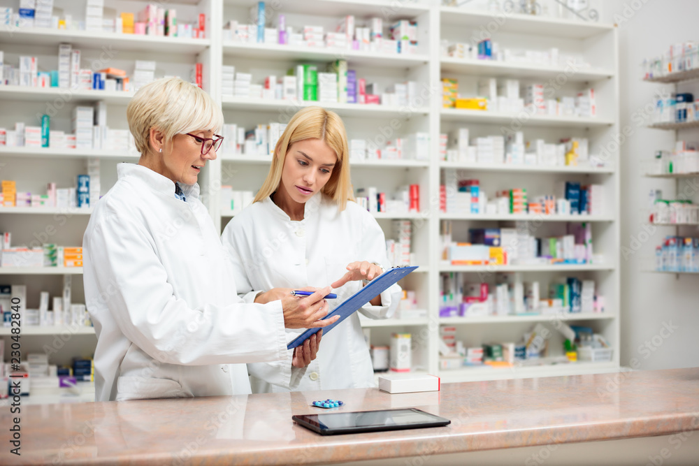 Mature and young female pharmacists working together, standing behind the counter and going over a checklist on a clipboard. Medicine and healthcare concept