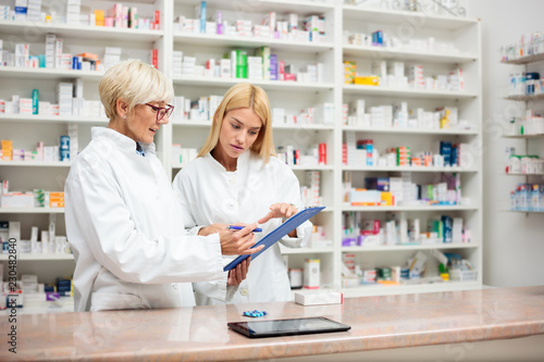 Mature and young female pharmacists working together, standing behind the counter and going over a checklist on a clipboard. Medicine and healthcare concept photo
