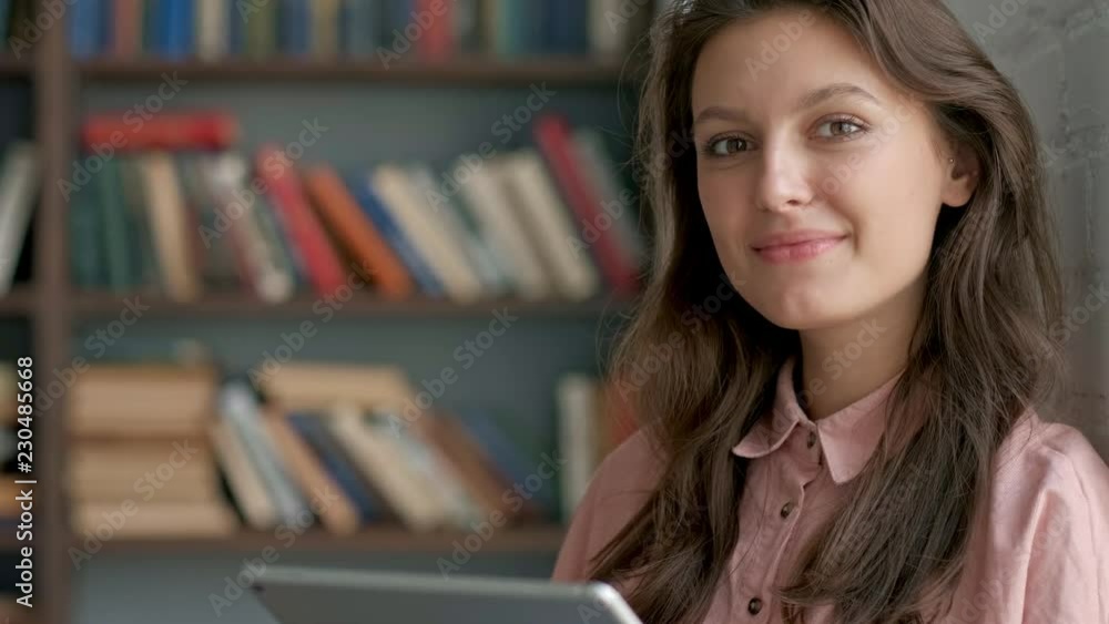 Close up of the curly young woman in glasses working on the tablet device and smiling to the camera at the library passage. Portrait. Indoors. Look at camera Close up of the curly young woman in glasses working on the tablet device and smiling to the camera at the library passage. Portrait. Indoors. Look at camera